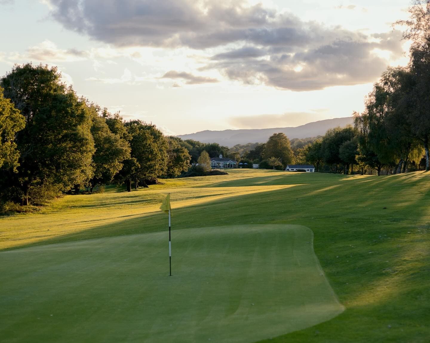 A green and flag at Glynneath Golf Club in evening light, with the fairway and clubhouse beyond